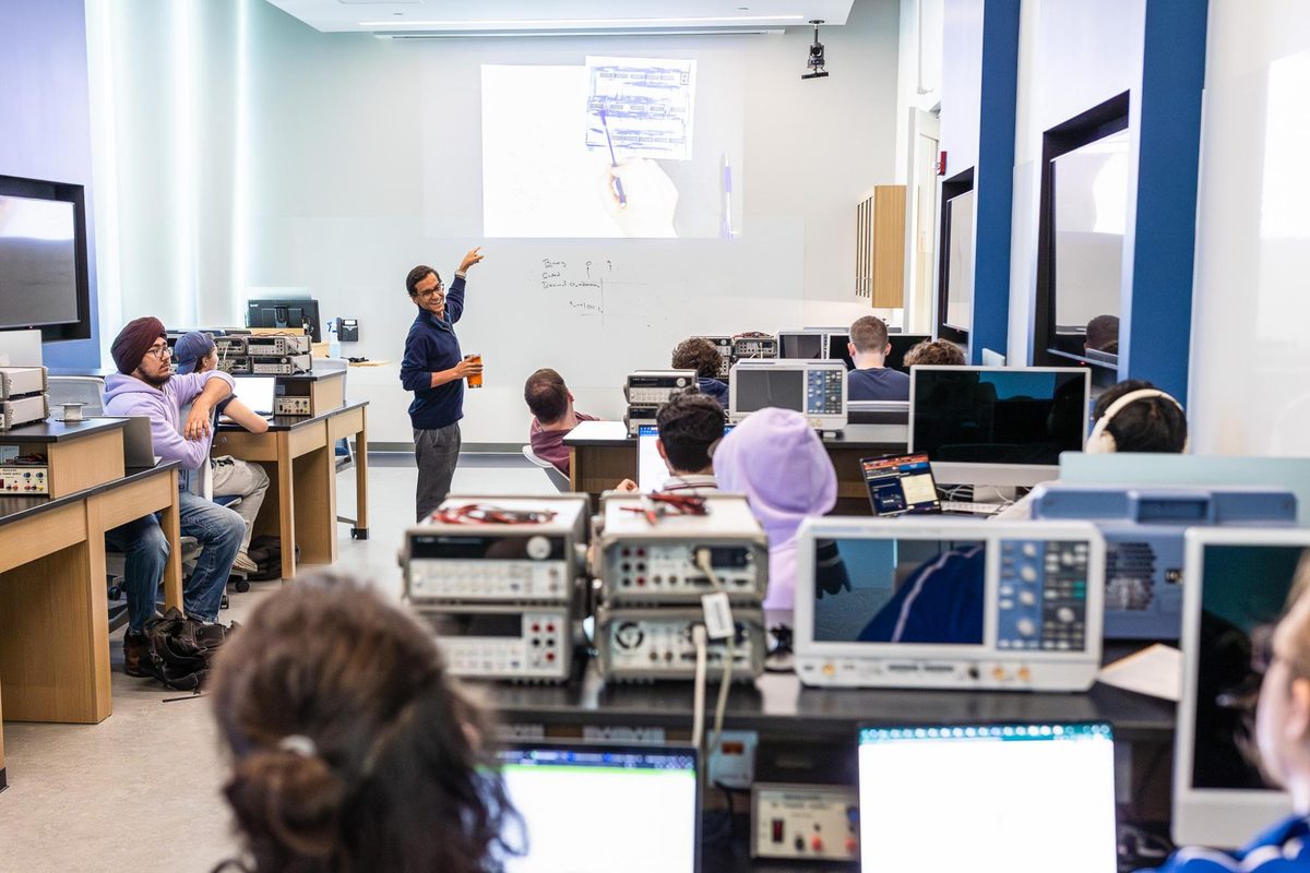 An instructor teaching at the front of the Architecture Lab while students work at benches with oscilloscopes and other test instruments